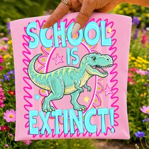 Op de afbeelding: Een lichtroze stoffen item met een centrale illustratie van een groene Tyrannosaurus Rex dinosaurus. De tekst "SCHOOL IS EXTINCT!" is gedrukt in grote, lichtblauwe letters. Het ontwerp bevat roze en gele steraccenten en een roze rand.