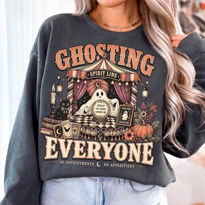 Pode incluir: Uma camisola de gola redonda cinzento-carv&atilde;o com uma impress&atilde;o gr&aacute;fica de um fantasma com turbante atr&aacute;s de uma bola de cristal, com o texto GHOSTING EVERYONE, NO APPOINTMENTS, NO APPARITIONS, SPIRIT LINE, HELLO? YOU THERE? ANSWER ME e AVAILABLE GHOSTING.