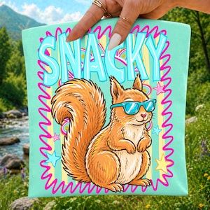 May include: A light mint green tote bag featuring a graphic illustration of a brown squirrel wearing bright blue sunglasses. The word SNACKY is printed in large blue letters above the squirrel, surrounded by a pink wavy border and yellow stars.