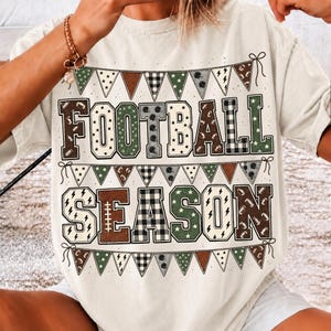 May include: A cream-colored t-shirt featuring the text FOOTBALL SEASON in large block letters. The letters are filled with patterns including brown, green, and black-and-white plaid. Bunting flags with football motifs border the text.