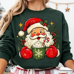 May include: A dark green long-sleeve sweatshirt featuring a graphic of Santa Claus wearing a red hat and red mittens, winking while holding a green mug filled with whipped cream and a candy cane, surrounded by holly leaves, berries, and stars.