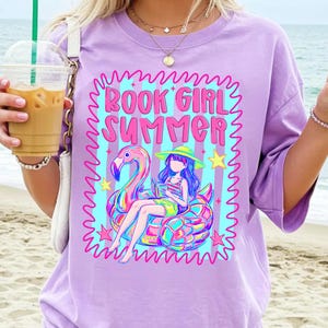 May include: A light purple t-shirt featuring a graphic print with the text BOOK GIRL SUMMER in pink. The design includes a person wearing a hat and reading a book while sitting on a colourful flamingo pool float, surrounded by stars and stripes.