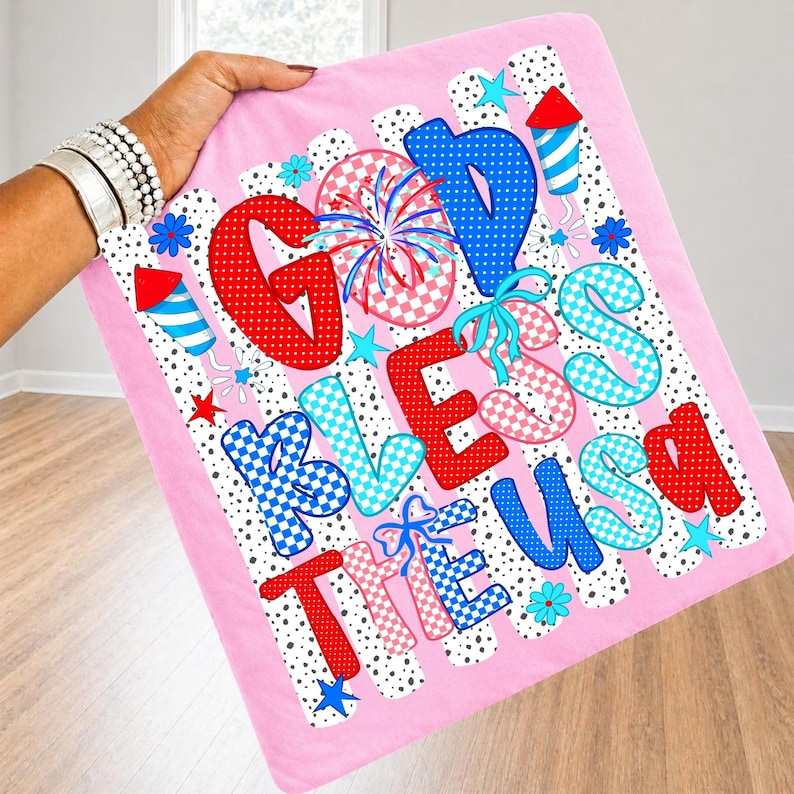 K&ouml;nnte beinhalten: Ein rosa Stoffquadrat mit dem Text GOD BLESS THE USA in rot, blau und wei&szlig; gemusterten Buchstaben. Das Design enth&auml;lt Illustrationen von Feuerwerk, Raketen und Sternen in patriotischen Farben vor einem schwarz-wei&szlig; gepunkteten Hintergrund.