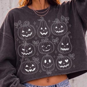 May include: A charcoal grey cropped sweatshirt featuring a white line-art graphic of nine various jack-o'-lanterns with different facial expressions, interspersed with small star shapes and decorative bows across the front of the garment.