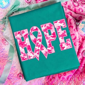 Peut inclure: Un t-shirt sarcelle avec le mot HOPE en grandes lettres majuscules. La lettre O est remplac&eacute;e par un ruban de sensibilisation rose. Les lettres pr&eacute;sentent un motif camouflage rose et blanc avec une texture us&eacute;e sur tout le graphisme.