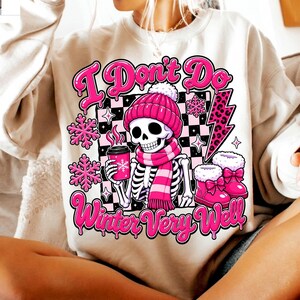 May include: A beige crewneck sweatshirt featuring a graphic of a skeleton wearing a pink beanie and scarf, holding a pink coffee cup. The design includes pink snowflakes, a leopard print lightning bolt, and the text I Don't Do Winter Very Well.