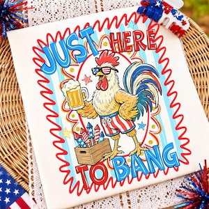 Op de afbeelding: Een wit t-shirt met een afbeelding van een haan met een zonnebril en een broek in de kleuren van de Amerikaanse vlag, die een bierpul vasthoudt. De haan staat naast een houten kist met vuurwerk. De tekst luidt JUST HERE TO BANG.