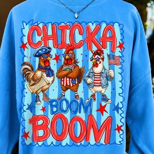 May include: A light blue sweatshirt featuring a graphic of three chickens wearing patriotic accessories and sunglasses. The text reads CHICKA BOOM BOOM. The chickens are depicted with American flag elements, including a flag, hat, and vest, set against a blue striped background with red and blue stars.