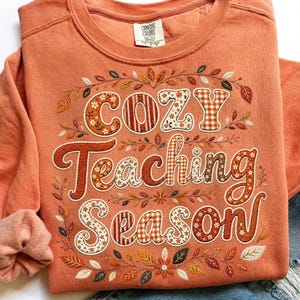 May include: A burnt orange crewneck sweatshirt featuring the text "Cozy Teaching Season" in a decorative font. The letters are filled with various patterns including floral, plaid, and solid colors, surrounded by an embroidered autumn leaf motif.