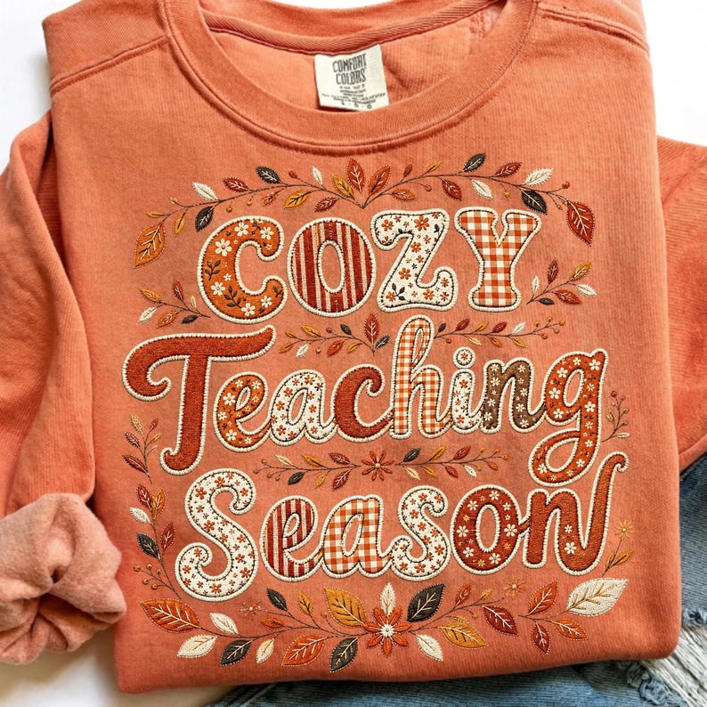 May include: A burnt orange crewneck sweatshirt featuring the text "Cozy Teaching Season" in a decorative font. The letters are filled with various patterns including floral, plaid, and solid colors, surrounded by an embroidered autumn leaf motif.