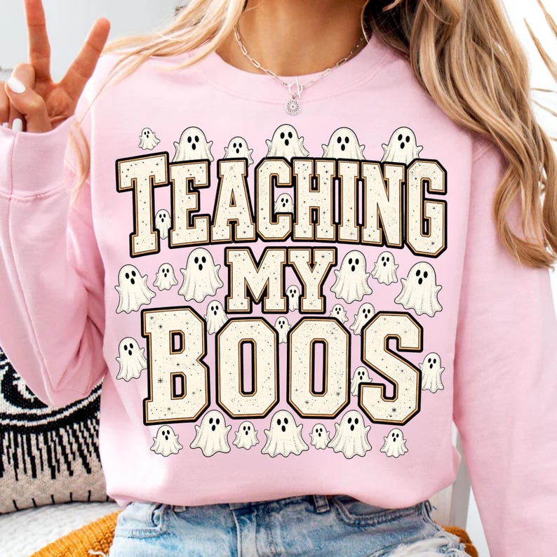 May include: A light pink crewneck sweatshirt featuring the text "TEACHING MY BOOS" in large, blocky, cream-colored letters with a dark outline. The design is surrounded by numerous small, white cartoon ghost illustrations.