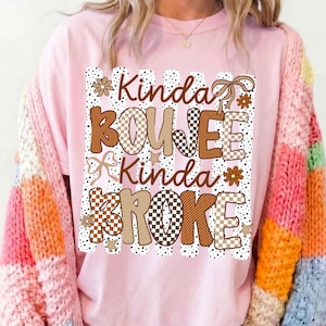 Pode incluir: Uma t-shirt rosa claro com uma estampa gr&aacute;fica com o texto Kinda BOUJEE Kinda BROKE. O design inclui padr&otilde;es xadrez castanhos e beges, riscas com bolinhas, pequenas estrelas, flores e la&ccedil;os de fita numa paleta de cores castanhas.