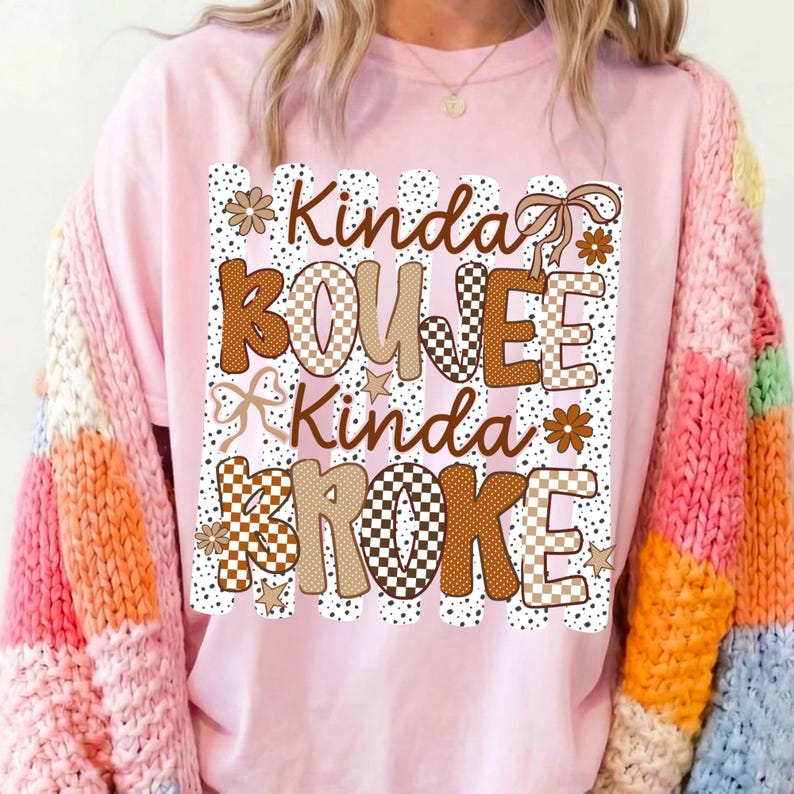 Pode incluir: Uma t-shirt rosa claro com uma estampa gr&aacute;fica com o texto Kinda BOUJEE Kinda BROKE. O design inclui padr&otilde;es xadrez castanhos e beges, riscas com bolinhas, pequenas estrelas, flores e la&ccedil;os de fita numa paleta de cores castanhas.