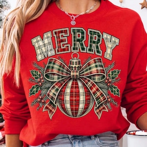 Puede incluir: Una sudadera roja de cuello redondo con un estampado gr&aacute;fico de la palabra MERRY en un patr&oacute;n de cuadros verdes y beige. Debajo del texto hay un gran adorno navide&ntilde;o de cuadros rojos y verdes con un lazo a juego y hojas de acebo.