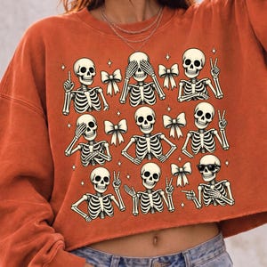 May include: A burnt orange long-sleeve sweatshirt featuring a graphic print of nine skeletons in various poses, including some making peace signs, covering their eyes, or wearing sunglasses, interspersed with small white bows and star shapes.
