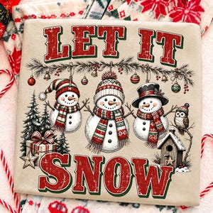 Pu&ograve; includere: Una t-shirt beige con la scritta rossa e verde LET IT SNOW. La grafica include tre pupazzi di neve con sciarpe e cappelli, ornamenti appesi, alberi di pino, un pacco regalo, una casetta per uccelli e un gufo in stile festivo.