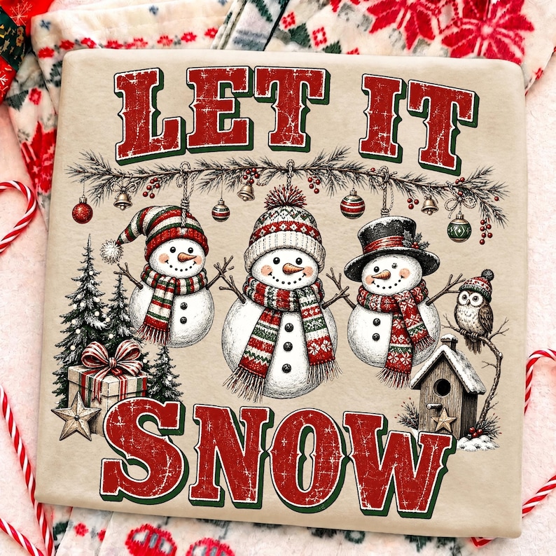 Pu&ograve; includere: Una t-shirt beige con la scritta rossa e verde LET IT SNOW. La grafica include tre pupazzi di neve con sciarpe e cappelli, ornamenti appesi, alberi di pino, un pacco regalo, una casetta per uccelli e un gufo in stile festivo.