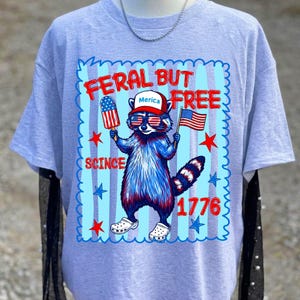 May include: A light grey t-shirt featuring a graphic of a raccoon wearing a red, white, and blue hat with the text Merica, holding a popsicle and an American flag. The text reads FERAL BUT FREE SCINCE 1776, surrounded by red and blue stars.