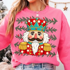 Puede incluir: Una sudadera de color rosa brillante con un gr&aacute;fico de un cascanueces que lleva una corona roja y turquesa, una chaqueta roja con charreteras doradas y una barba blanca. El dise&ntilde;o est&aacute; rodeado de ramas de pino verdes y bombillas rojas.