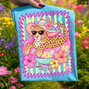 Könnte beinhalten: Ein hellblaues T-Shirt mit einem Cartoon-Leoparden, der eine Sonnenbrille und einen Sonnenhut trägt und in einem rosa aufblasbaren Ring liegt. Das Design umfasst Hibiskusblüten und einen wellenförmigen Rand.