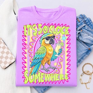 May include: A lilac-colored t-shirt featuring a cartoon parrot wearing sunglasses and holding a cocktail. The shirt has the text "IT'S 5 O'CLOCK SOMEWHERE" in green and yellow. The parrot is in an oval frame with a floral design.