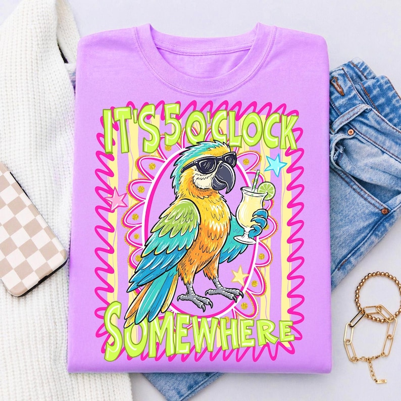 May include: A lilac-colored t-shirt featuring a cartoon parrot wearing sunglasses and holding a cocktail. The shirt has the text "IT'S 5 O'CLOCK SOMEWHERE" in green and yellow. The parrot is in an oval frame with a floral design.