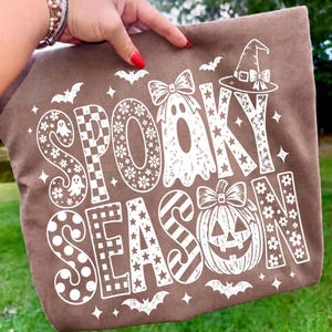 May include: A brown fabric item featuring a white graphic print with the text SPOOKY SEASON. The design includes a ghost wearing a bow, a pumpkin with a bow, stars, flowers, checkered patterns, polka dots, stripes, and small bat silhouettes.