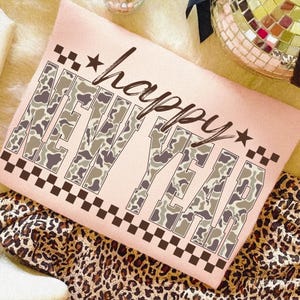 May include: A light pink cushion featuring the words "happy NEW YEAR" in a camouflage design. The cushion has a black and brown chequered border and star accents. It rests on leopard print fabric.