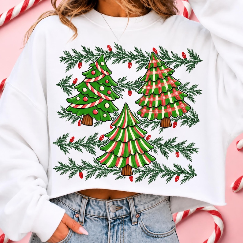 May include: A white long-sleeve cropped sweatshirt featuring a graphic print of three stylised Christmas trees. The trees are decorated with red and white stripes and plaid patterns, surrounded by horizontal rows of green pine branches and red lights.