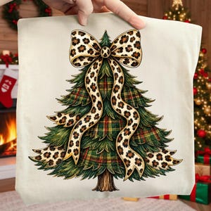 May include: A cream-colored fabric item featuring a graphic illustration of a Christmas tree. The tree has green branches with a red, green, and yellow plaid pattern and is topped with a large, flowing bow in a tan and black leopard print design.