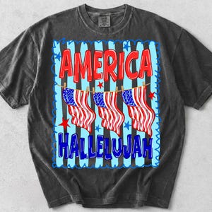 Puede incluir: Una camiseta gris carb&oacute;n de manga corta con un estampado gr&aacute;fico que incluye el texto AMERICA HALLELUJAH en rojo y azul. El dise&ntilde;o muestra tres banderas estadounidenses colgadas en un tendedero sobre rayas de color azul claro y negro.