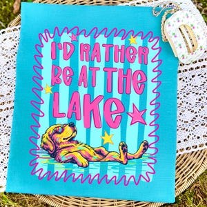 May include: A turquoise t-shirt featuring a graphic of a dog wearing sunglasses, lying down. The text reads "I'D RATHER BE AT THE LAKE" in pink letters. The design includes a pink squiggly border, yellow stars, and a light blue striped background.