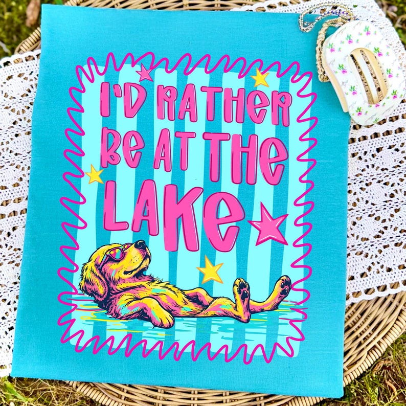 May include: A turquoise t-shirt featuring a graphic of a dog wearing sunglasses, lying down. The text reads "I'D RATHER BE AT THE LAKE" in pink letters. The design includes a pink squiggly border, yellow stars, and a light blue striped background.