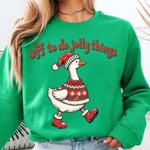 May include: A bright green crewneck sweatshirt featuring a graphic of a white goose wearing a red and green patterned sweater, a matching Santa hat, and red boots. The text "off to do jolly things" is printed in red above the goose illustration.