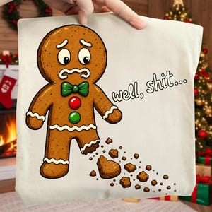May include: A cream-coloured t-shirt featuring a cartoon gingerbread man with a worried expression, a green bow tie, and red and green buttons. The gingerbread man has a missing foot with scattered crumbs and the text well, shit...