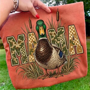 May include: A coral-colored fabric featuring a graphic of a mallard duck with a green head and yellow beak. The word MAMA is printed in large block letters with a camouflage and checkerboard pattern, surrounded by illustrations of tall grass.