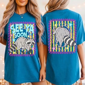 Op de afbeelding: Een blauwgroen t-shirt met een afbeelding van een wasbeer en de tekst "SEE YA SOON" op de voorkant, en een bijpassende afbeelding van een wasbeer op de achterkant. Het ontwerp bevat gele verticale strepen, roze randen en sterren.