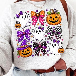 May include: A light grey sweatshirt featuring a graphic print of three white ghosts, two orange jack-o'-lanterns, and five decorative bows in various patterns including polka dots and checkers, accented with small purple and orange stars.