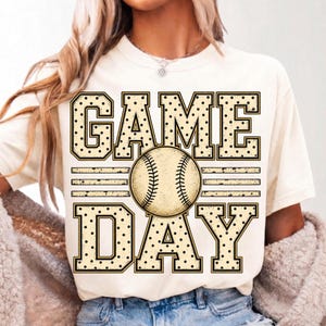 May include: A cream-coloured short-sleeve t-shirt featuring a graphic print with the text GAME DAY in large, block-style letters with a black polka dot pattern. A detailed illustration of a baseball is positioned between the words.
