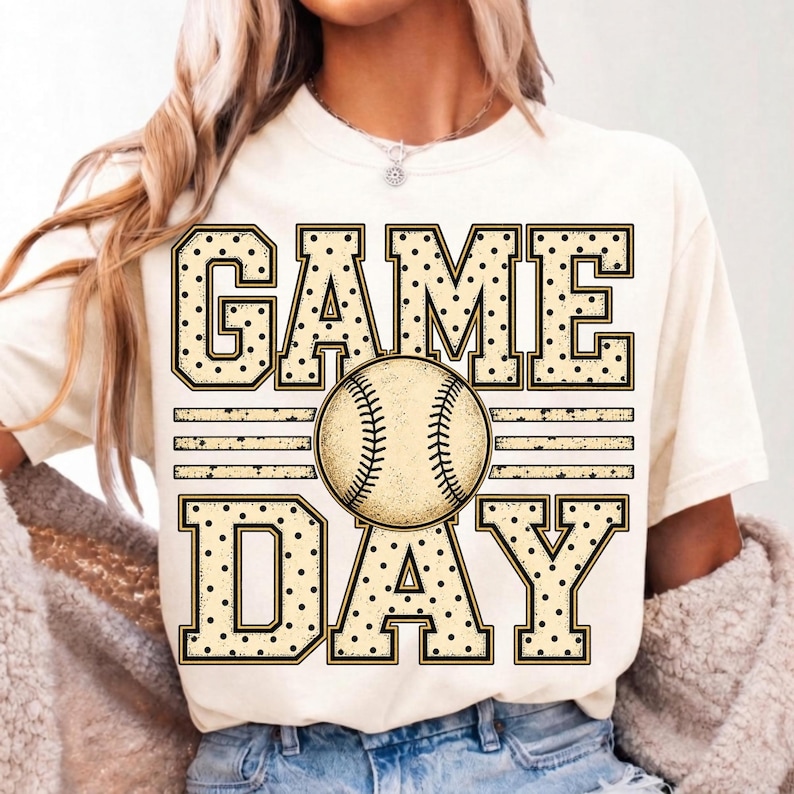 May include: A cream-coloured short-sleeve t-shirt featuring a graphic print with the text GAME DAY in large, block-style letters with a black polka dot pattern. A detailed illustration of a baseball is positioned between the words.