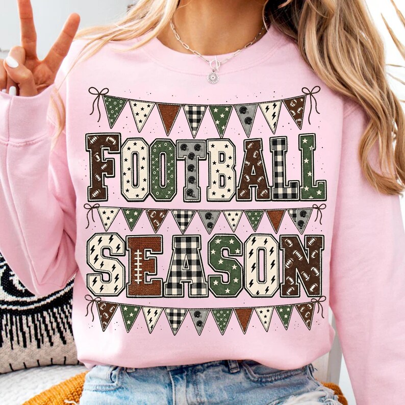 May include: A light pink crewneck sweatshirt featuring the text FOOTBALL SEASON in large block letters. The letters are filled with various patterns including plaid, stars, and footballs. The design is framed by rows of triangular bunting flags.