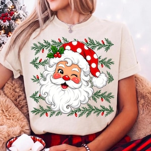 Puede incluir: Una camiseta de color crema con un gr&aacute;fico de un Santa Claus gui&ntilde;ando un ojo y llevando un gorro rojo con lunares blancos. El dise&ntilde;o incluye patrones de ramas de pino verdes y peque&ntilde;as bombillas rojas alrededor de la ilustraci&oacute;n.