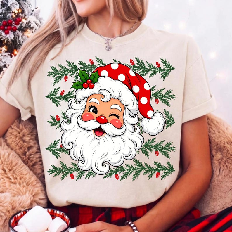 Puede incluir: Una camiseta de color crema con un gr&aacute;fico de un Santa Claus gui&ntilde;ando un ojo y llevando un gorro rojo con lunares blancos. El dise&ntilde;o incluye patrones de ramas de pino verdes y peque&ntilde;as bombillas rojas alrededor de la ilustraci&oacute;n.