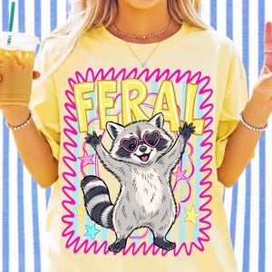 以下が含まれることがあります： 淡い黄色のTシャツで、ピンクのハート型サングラスをかけた灰色のラクーンのグラフィックが描かれています。ラクーンは両手を上げており、その上には大きな黄色の文字で「FERAL」とプリントされ、全体が鮮やかなピンク色の波打つ枠で囲まれています。