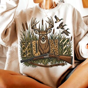 May include: A beige crewneck sweatshirt featuring a graphic print of a deer head, three flying ducks, and cattails. The word HUNT is printed in large block letters with camouflage and leopard patterns. A double-barrelled shotgun is positioned below.