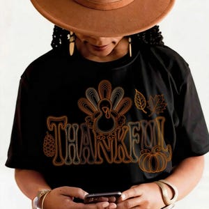 May include: A black short-sleeved t-shirt featuring a graphic design in orange and gold outlines. The design includes the word THANKFUL, a turkey, a pinecone, two leaves, and a pumpkin. The shirt is worn with a wide-brimmed brown hat.