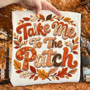 May include: A white t-shirt featuring the embroidered text Take Me To The Patch in orange, brown, and cream colors. The letters have various patterns including gingham, floral, and solid textures, surrounded by a wreath of autumn leaves.