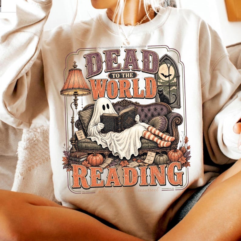 May include: A beige crewneck sweatshirt featuring a graphic print of a ghost sitting on a sofa reading a book. The text reads DEAD TO THE WORLD READING. The design includes a floor lamp, a window with a bat and cat, pumpkins, and autumn leaves.