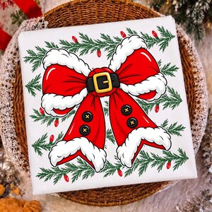 Puede incluir: Una camiseta blanca con un gr&aacute;fico de un gran lazo rojo con borde de piel blanca, un cintur&oacute;n negro con hebilla dorada y botones negros. El dise&ntilde;o est&aacute; rodeado por ramas de pino verdes y peque&ntilde;as luces navide&ntilde;as rojas.