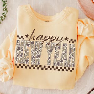 May include: A light yellow sweatshirt with the words "happy NEW YEAR" in a camouflage pattern. The word "happy" is in cursive, and the words are framed by stars and a black and white checkered pattern.